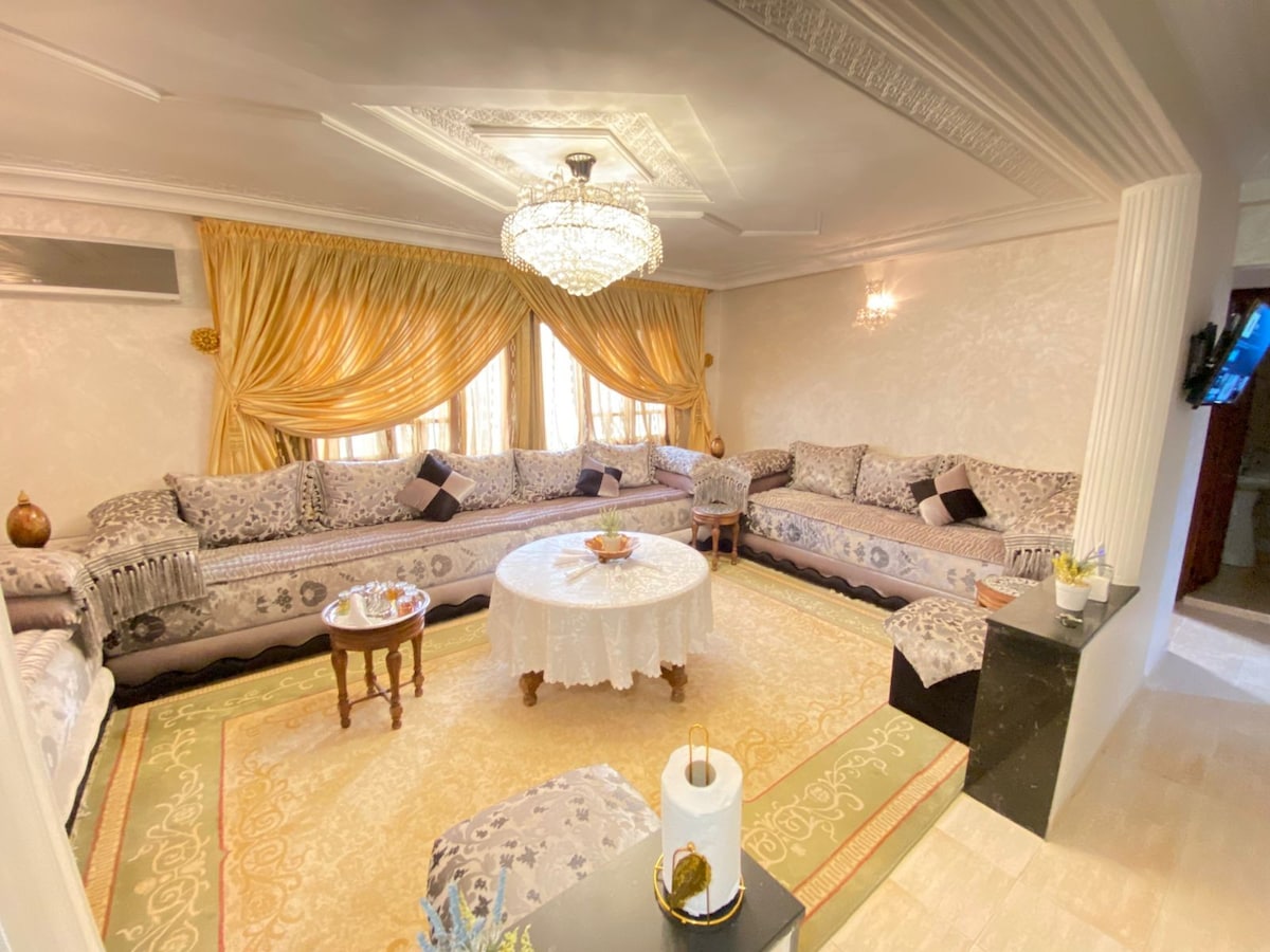 A spacious living area is adorned with elegant gold drapes and a chandelier. Multiple cushioned sofas surround a low circular table, while a patterned area rug adds warmth to the decor. Natural light enters through large windows, enhancing the inviting atmosphere of the room.