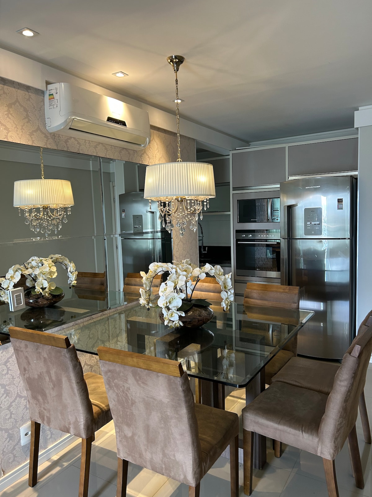 A dining area features a glass-top table surrounded by six upholstered chairs. A stylish chandelier hangs above, enhancing the ambiance. Mirrored walls reflect the space, while modern appliances are visible in the background, contributing to a functional and inviting dining experience.