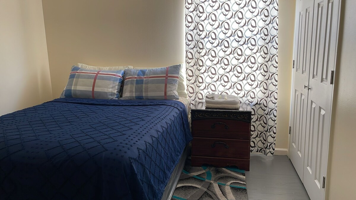 A spacious sleeping area features a large bed dressed in a patterned blue comforter. Two decorative pillows are placed against a stylish patterned headboard. A window with translucent drapes allows natural light, while a wooden chest serves as a nightstand, accompanied by neatly stacked towels.