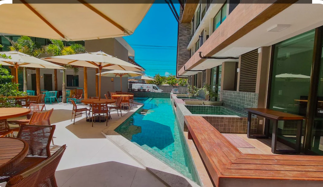 An outdoor area features a refreshing swimming pool flanked by lounge spaces. Tables and chairs provide seating around the poolside, with umbrellas offering shade. Lush greenery surrounds the space, enhancing the tranquil atmosphere of this leisure area.