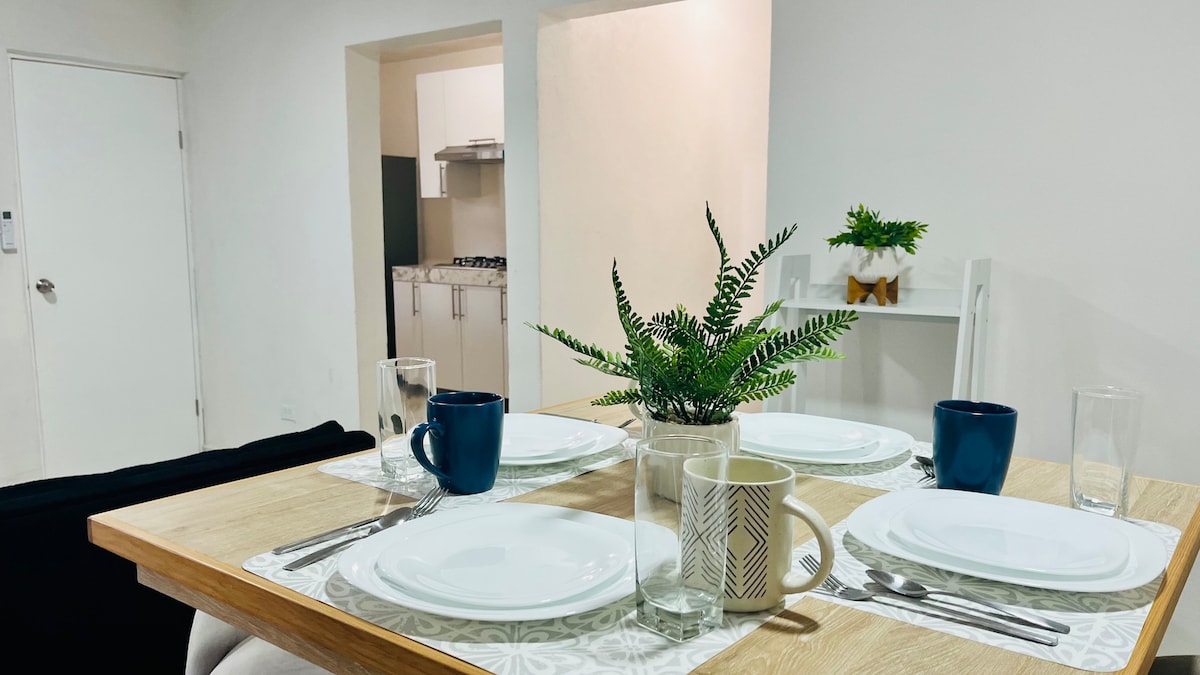 A dining table is set with four white plates, blue mugs, and glassware. A small potted plant provides a touch of greenery. The kitchen can be seen in the background, featuring modern appliances and a neutral color palette.