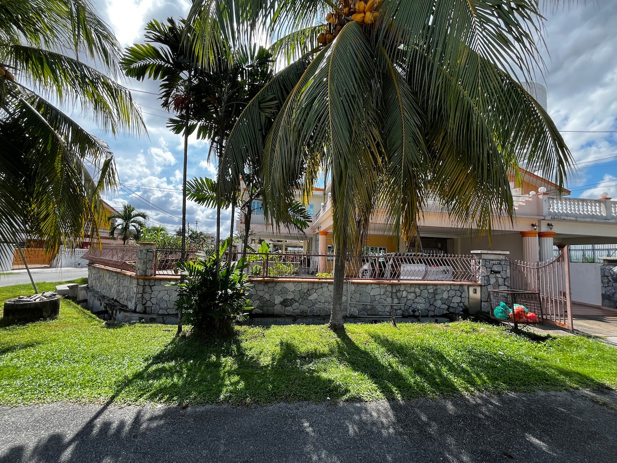 A spacious outdoor area is framed by lush palm trees and green grass. A stone wall and gated entrance delineate the property, with a glimpse of the house visible in the background. The sky is partly cloudy, suggesting a pleasant atmosphere.