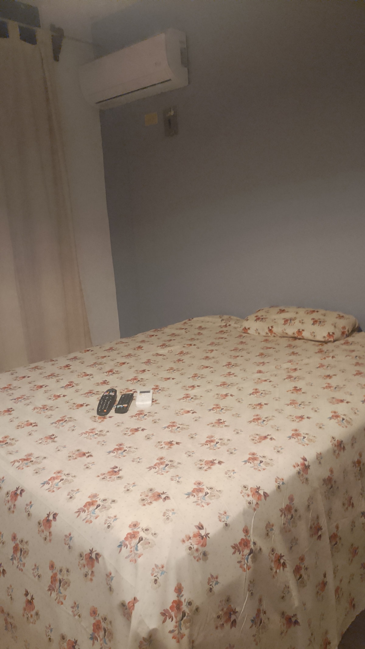A cozy sleeping area features a bed covered with a floral-patterned bedspread. A small pillow rests against the wall, and multiple remote controls are placed on the bed's surface. The room is lightly colored with a window partially covered by a curtain.