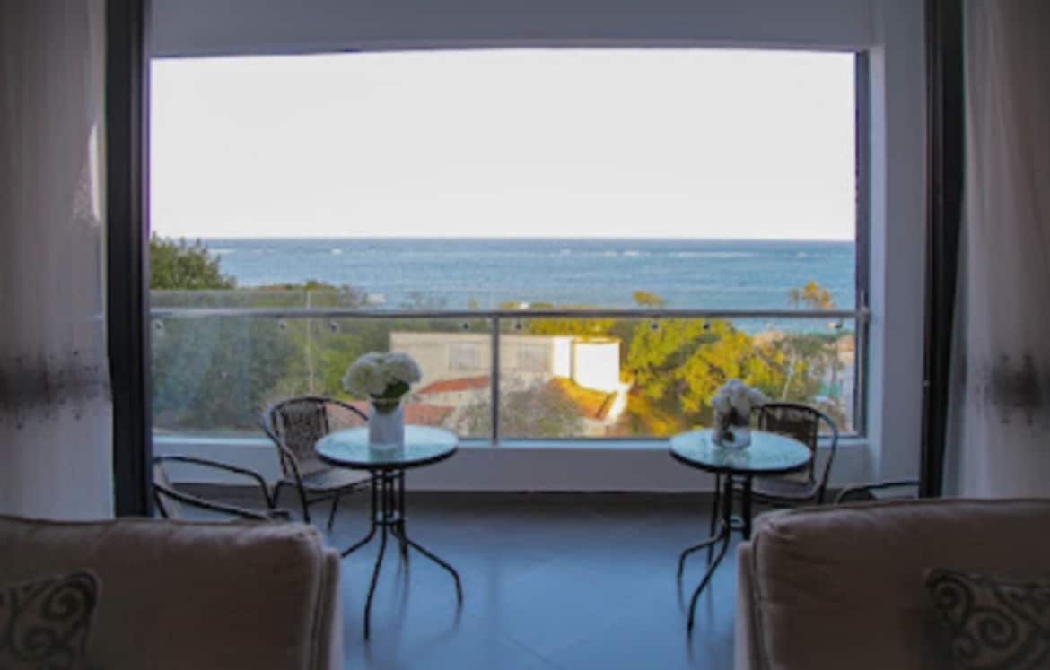A spacious balcony offers serene ocean views, featuring two round tables with chairs placed on either side. Bright sunlight illuminates the area, emphasizing the calm atmosphere created by the coastal scenery.