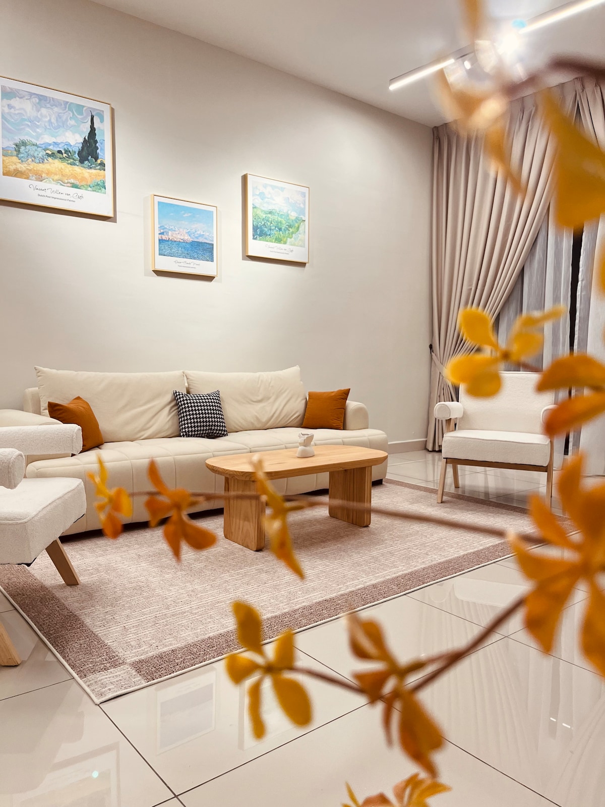 The living room features a light-colored sofa adorned with orange cushions, paired with two white armchairs. A coffee table sits centrally on a plush rug. Framed artworks hang on the walls, and soft curtains partially cover large windows, creating a bright and welcoming space.