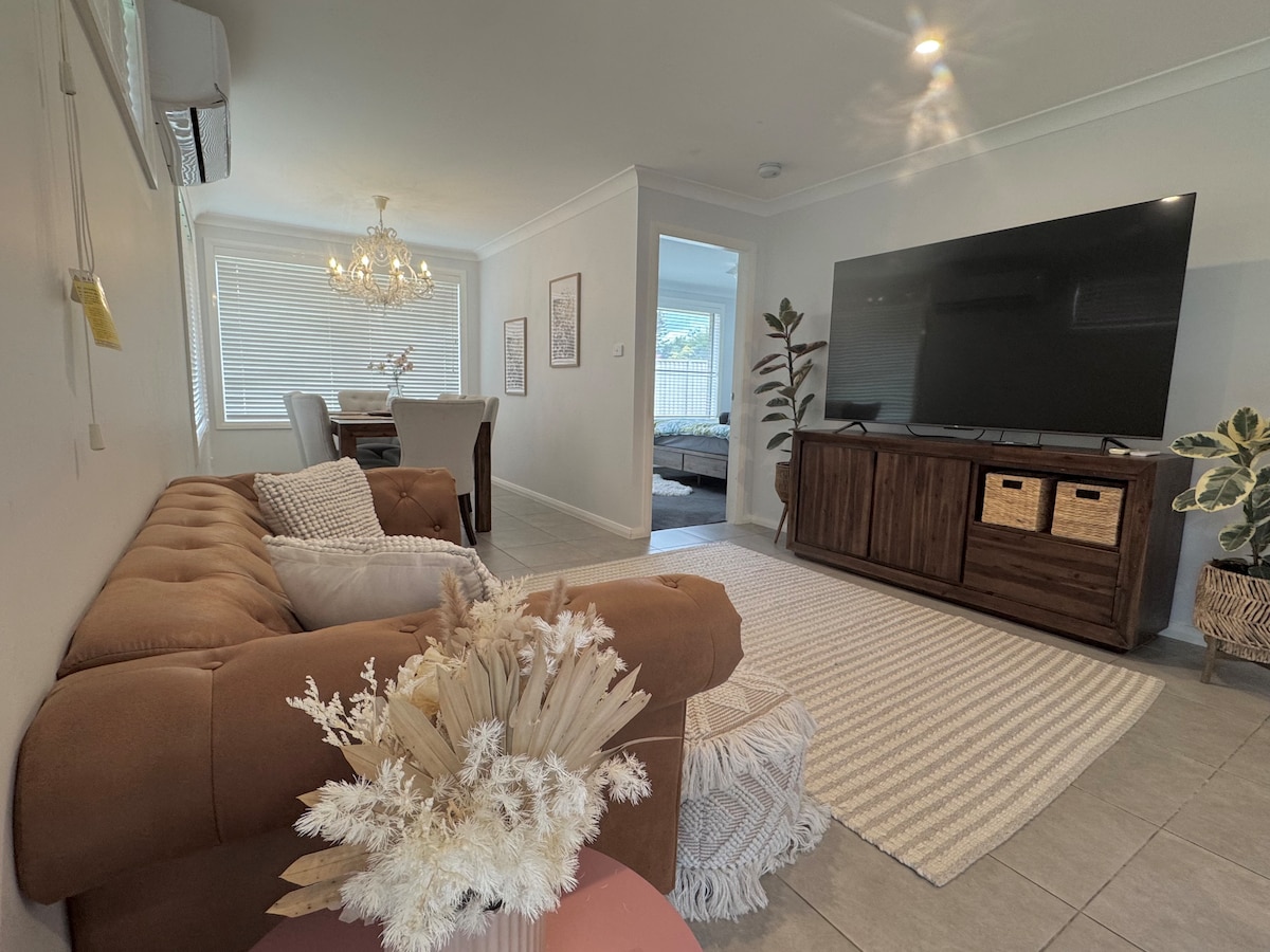 A comfortable living area is presented, featuring a brown sectional sofa with textured cushions. A patterned area rug complements a light-colored coffee table topped with dried floral arrangements. A large television is mounted on a wooden cabinet, with a dining area visible in the background.