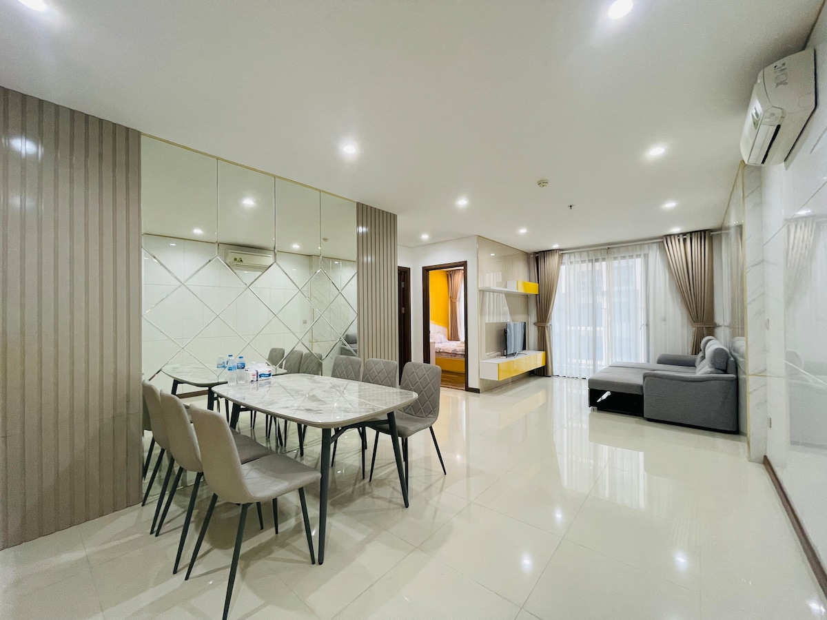 A spacious living area features a large marble dining table surrounded by six upholstered chairs. Soft lighting reflects off the glossy tile floors, while a cozy seating arrangement with a gray sofa adds comfort. A bright bedroom area is visible through glass doors, enhancing the open layout.