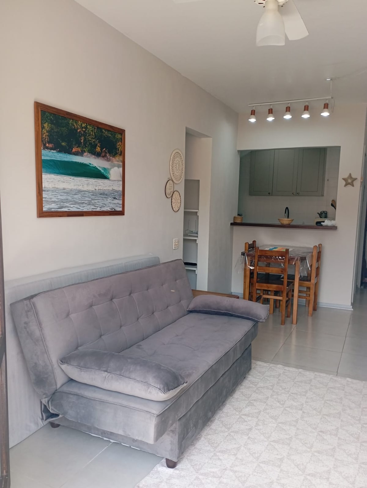 A comfortable living area features a gray sofa, positioned against a light wall adorned with a framed nature photograph. The open layout reveals a dining space and kitchen area in the background, illuminated by soft overhead lighting.