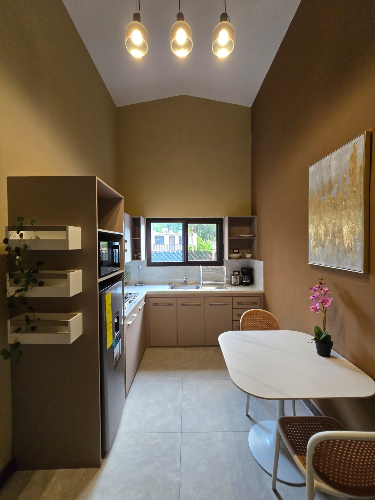 The kitchen features modern cabinetry with a sleek design and ample counter space. A window offers natural light, with a small dining table and two chairs positioned nearby. A potted plant and an orchid add a touch of decor to the space.