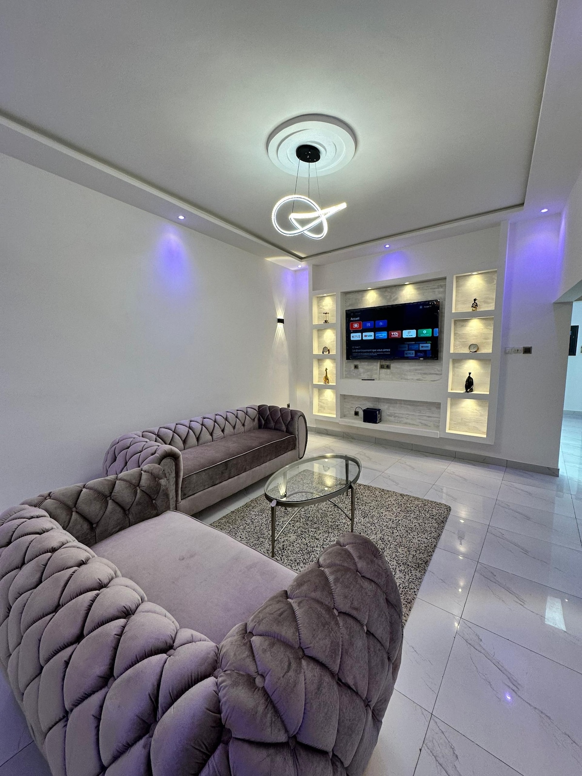 A modern living area features plush, tufted sofas in soft gray tones, positioned near a sleek glass table on a soft rug. Ambient lighting sets a relaxed mood, enhancing a wall-mounted entertainment center that is equipped with a television and decorative niches.