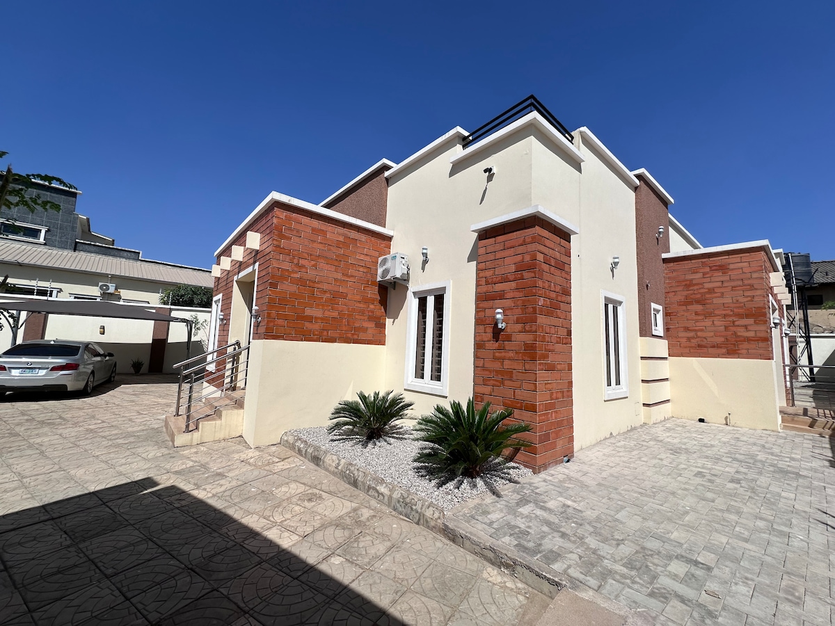 A modern home exterior features a combination of light-colored walls and warm brick accents. Lush plants are positioned alongside the entrance. The driveway, paved with stone, leads to an adjacent parking area, complemented by a clear blue sky above.
