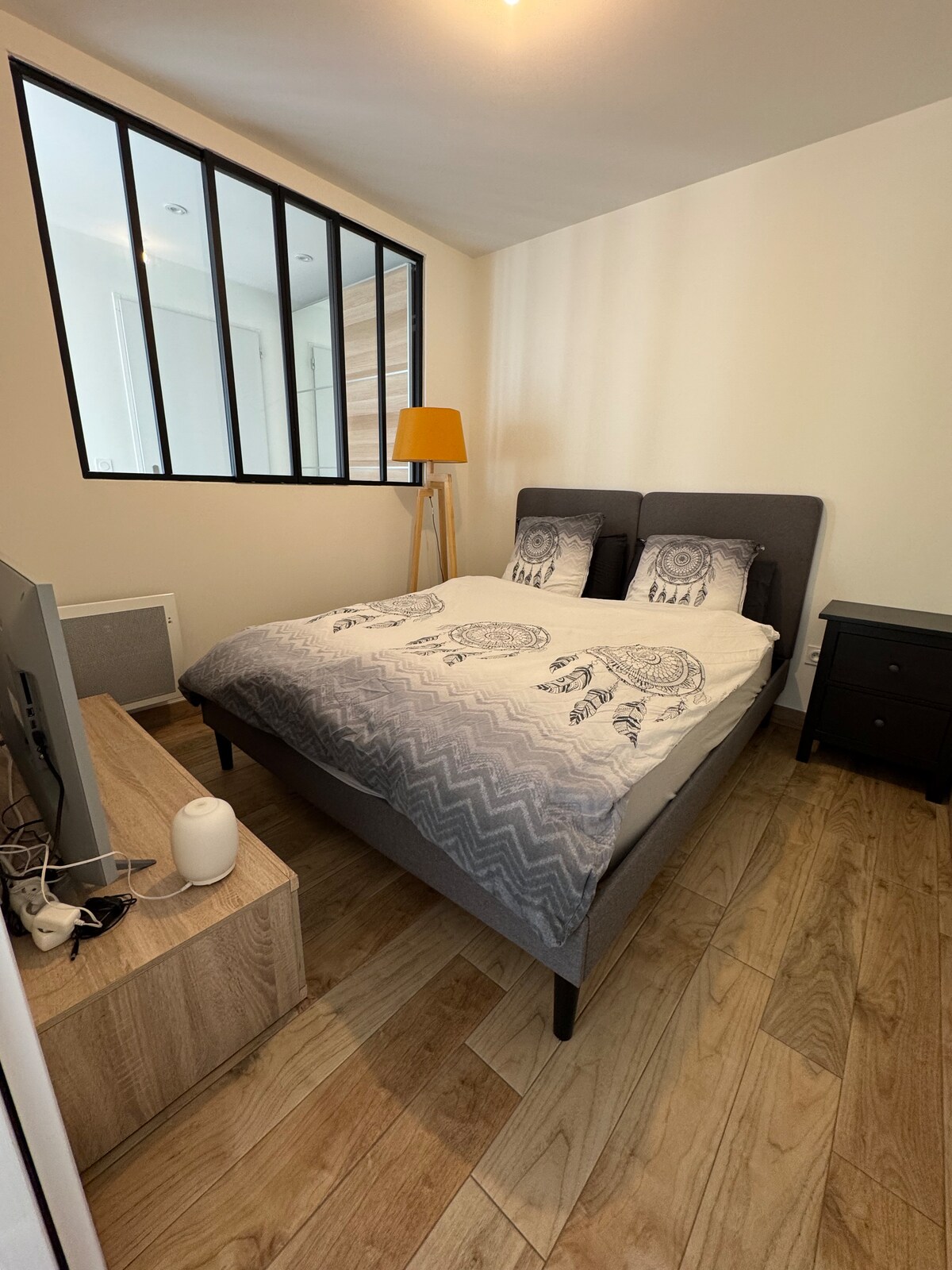 A contemporary bedroom features a spacious bed with patterned bedding and decorative pillows. A wooden nightstand and a black dresser are positioned beside the bed. A floor lamp adds illumination, while large windows allow natural light to fill the room.