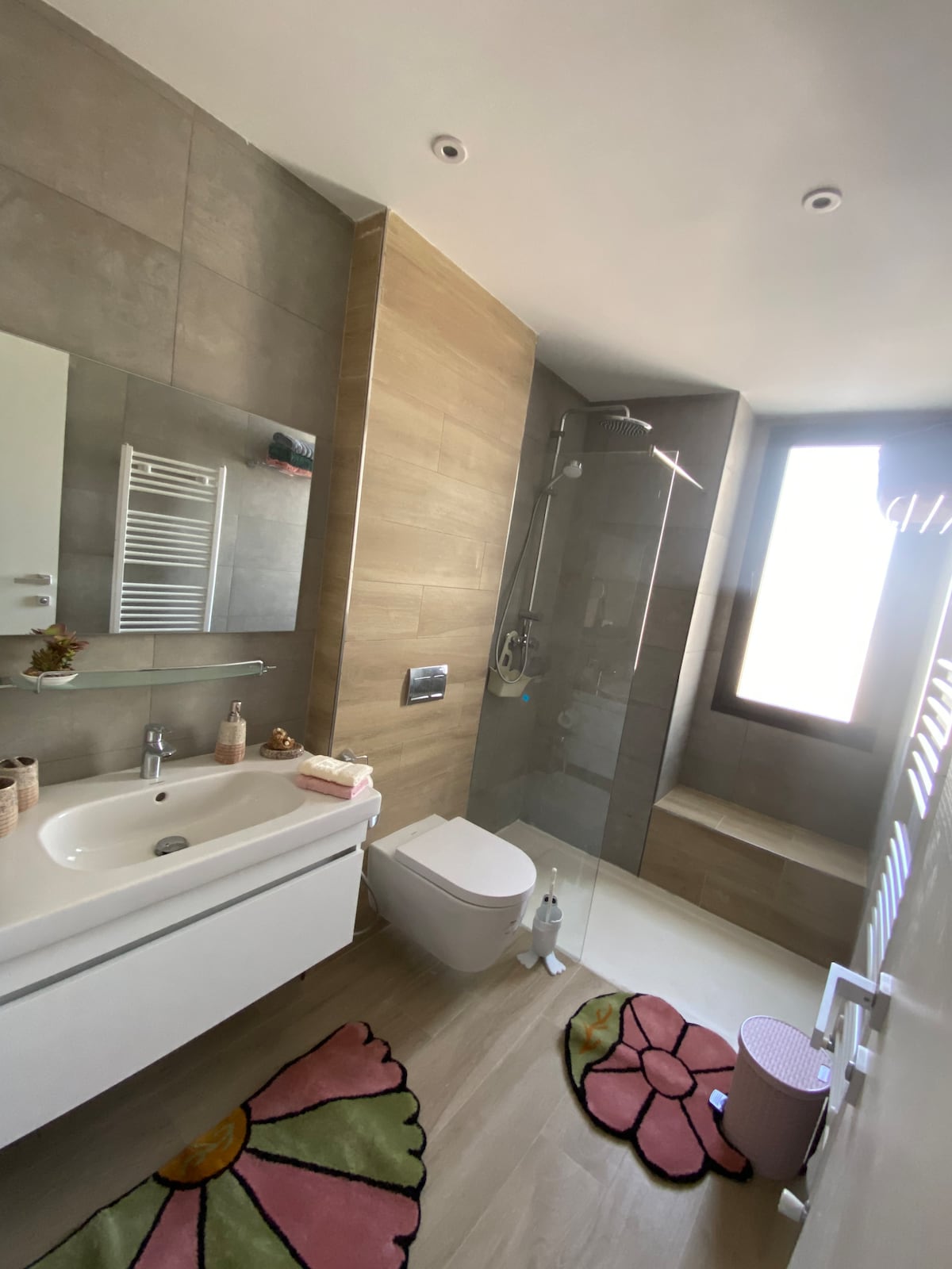 The bathroom features contemporary design elements including a spacious walk-in shower and modern fixtures. A white vanity with a clean countertop offers ample storage. Colorful floral rugs add a soft touch to the space, while natural light floods in through the tall window.