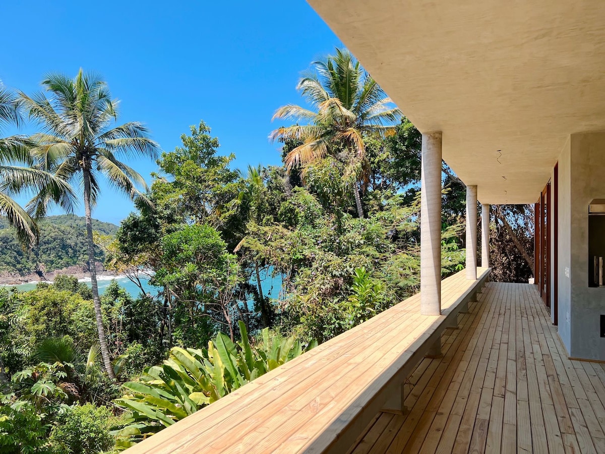 A broad wooden terrace is framed by palm trees and tropical foliage, offering views of a serene bay. The vertical columns provide structural support, while the open space invites natural light and fresh air, creating a connection with the surrounding landscape.