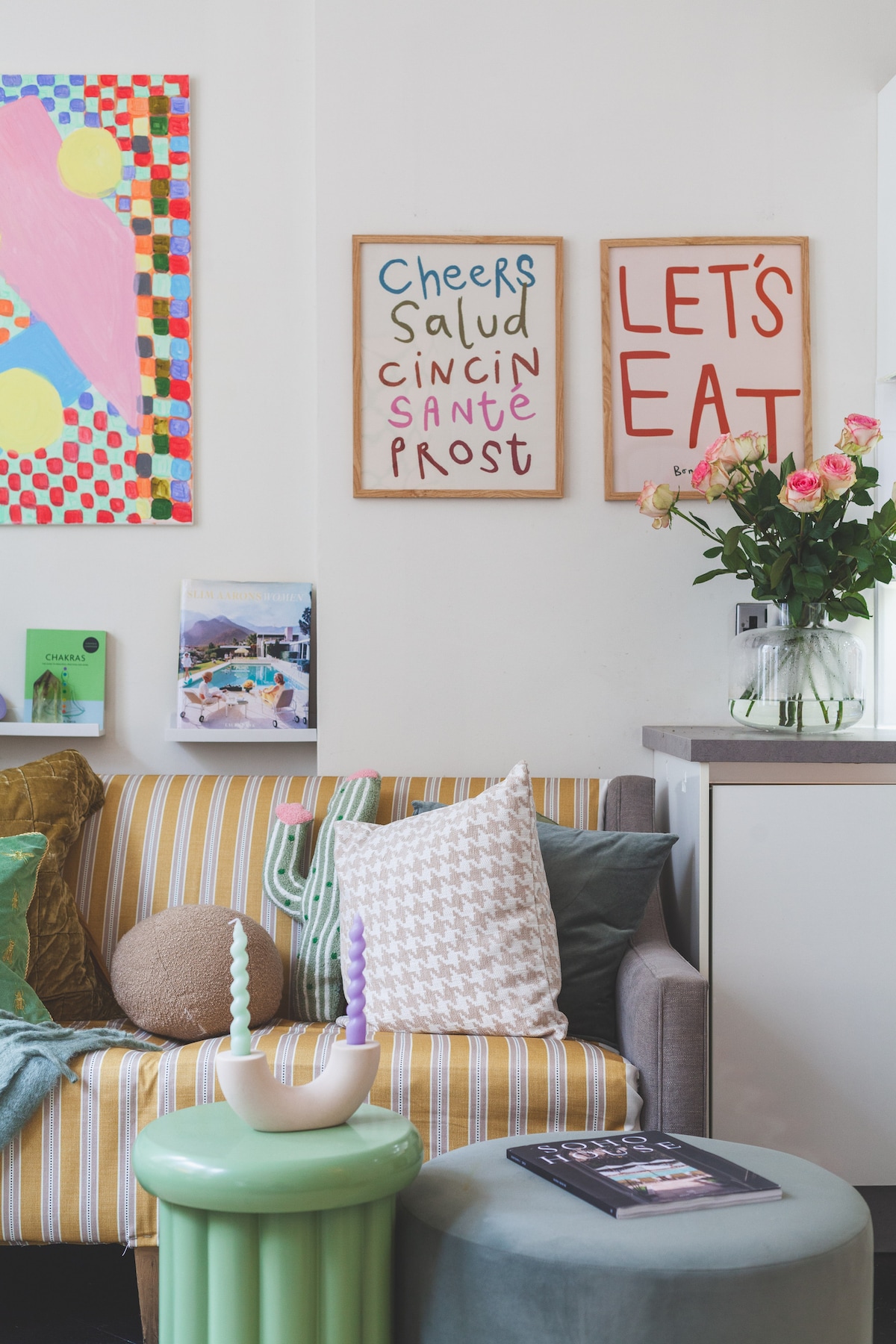A cozy corner features a striped sofa adorned with various cushions and a decorative throw. Two framed art pieces display cheerful phrases, while a small green table holds a vase of fresh flowers. A nearby magazine lays open on the surface.