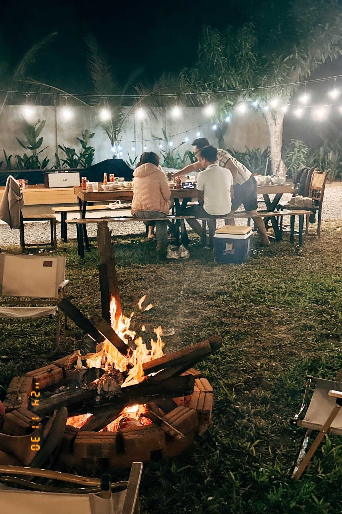 A cozy outdoor space is captured, featuring a warm fire pit at the center with flames gently flickering. A long wooden table is arranged with chairs, while string lights create a soft glow above. Two individuals are seated at the table, engaged in conversation.