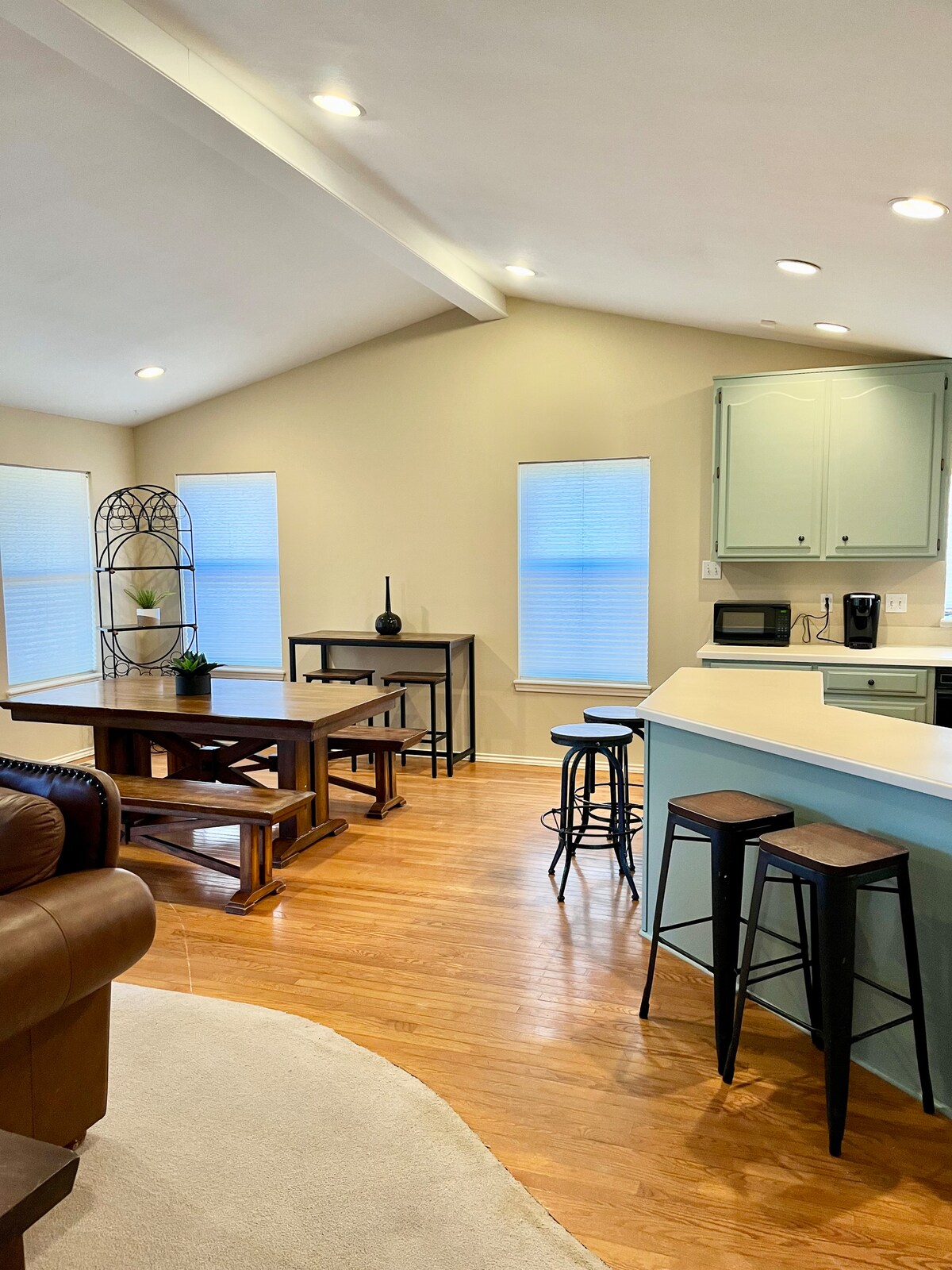 An open floor plan is visible, featuring a dining area with a large wooden table and several mixed-height stools at a kitchen counter. The space is illuminated by natural light from multiple windows, and a light blue cabinetry adds a subtle color accent.