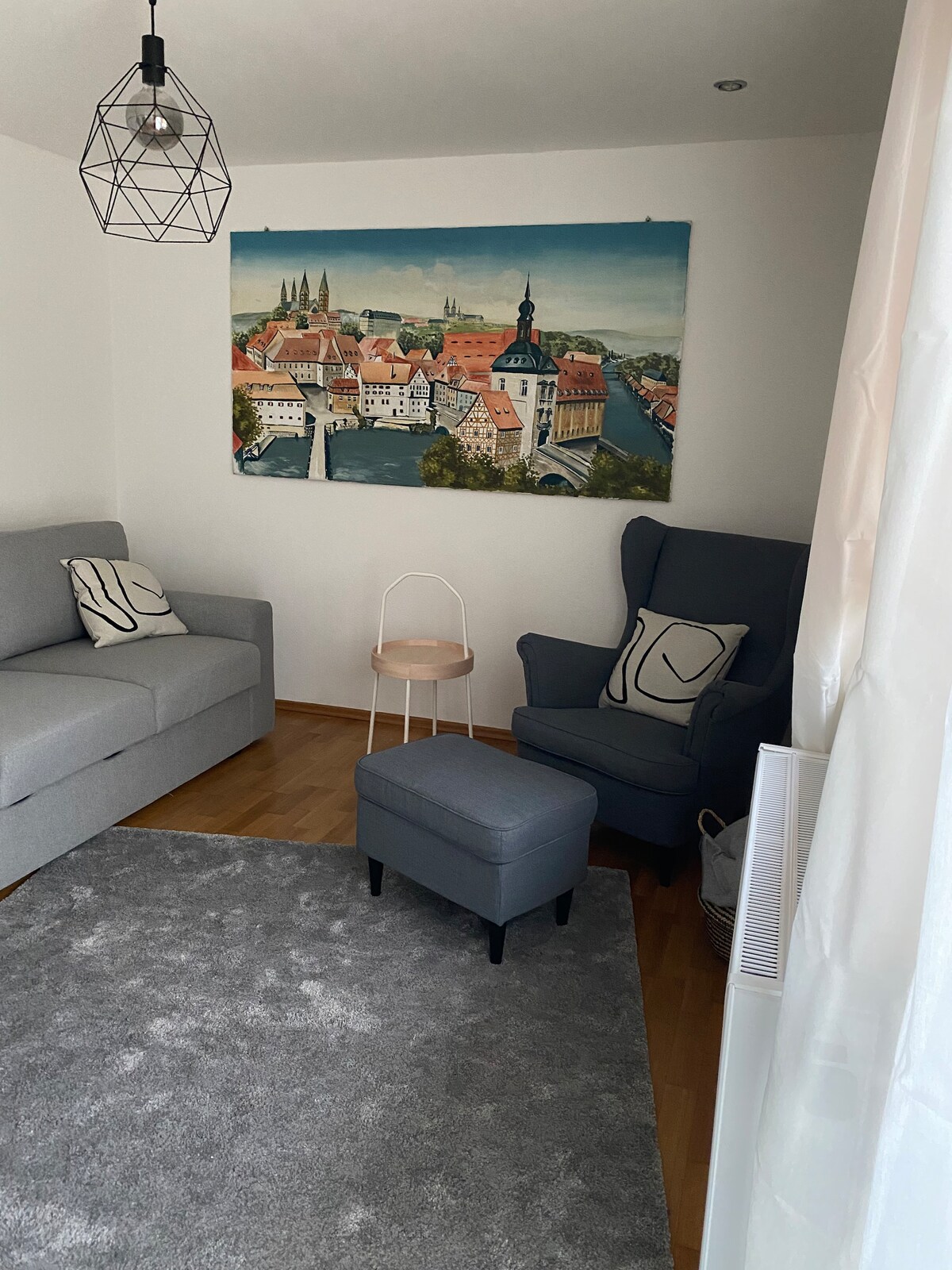 A living area features a light gray sofa and a dark blue armchair, accompanied by a small, round side table. A textured area rug lies beneath, while a large wall art piece depicting a town scene adds interest to the bright space.