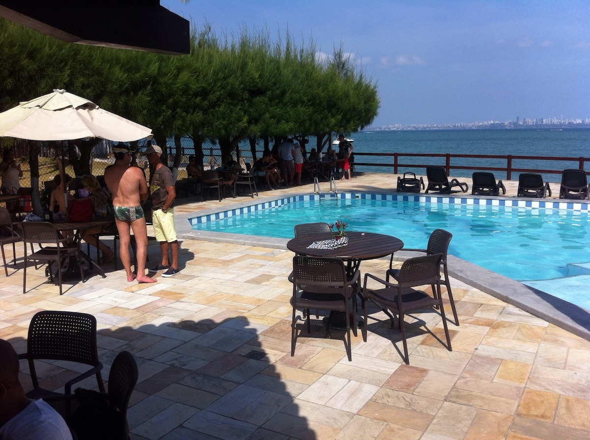 An outdoor pool area features a clear blue swimming pool surrounded by tiled flooring. Tables and chairs are arranged near the pool, with umbrellas providing shade. Lush greenery lines the backdrop, enhancing the ocean view in the distance.