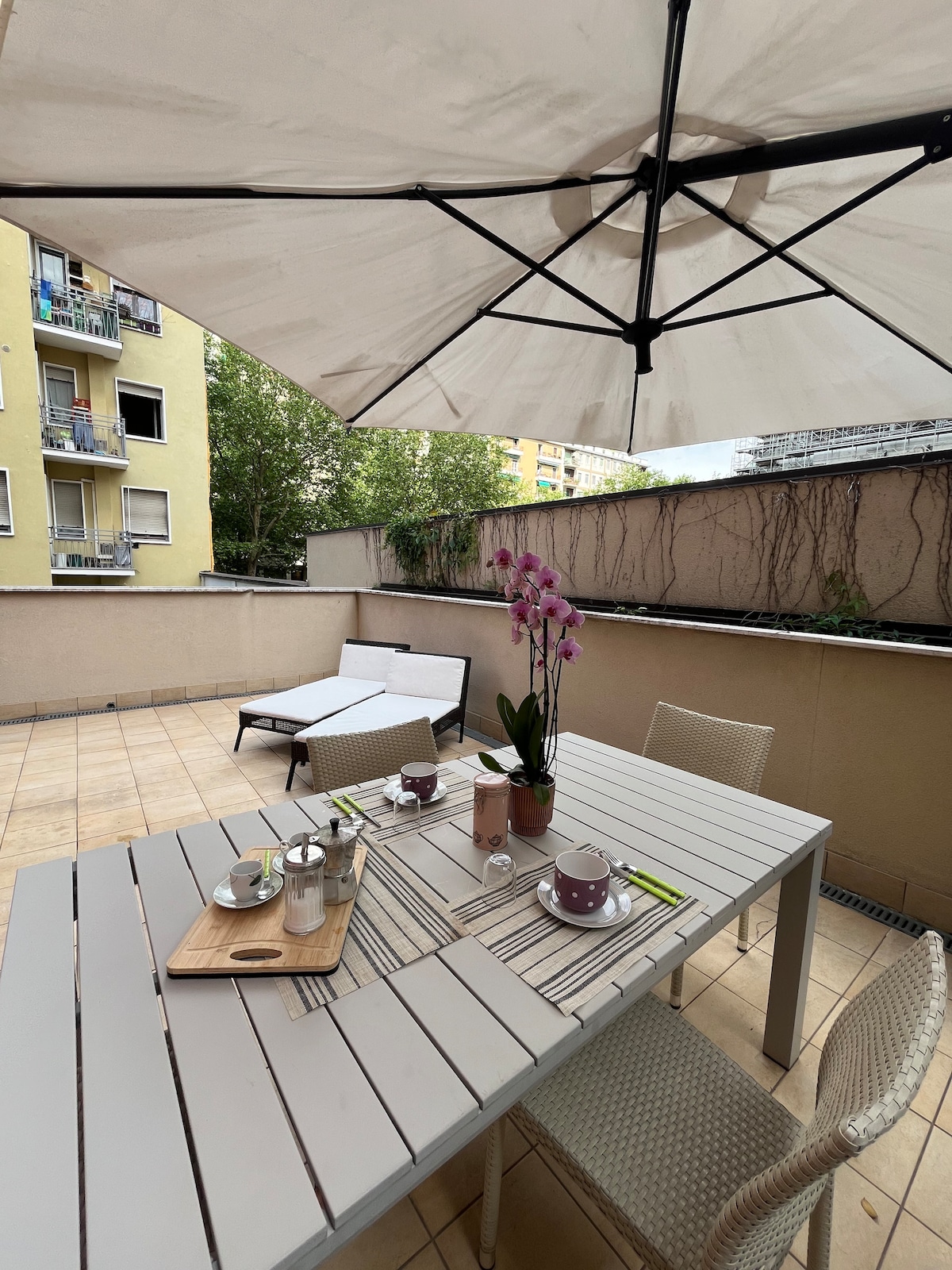 An outdoor terrace features a large umbrella providing shade over a dining table set for four. A potted orchid adds a touch of color, while a sun lounger is positioned to the side. Surrounding greenery offers a serene backdrop.