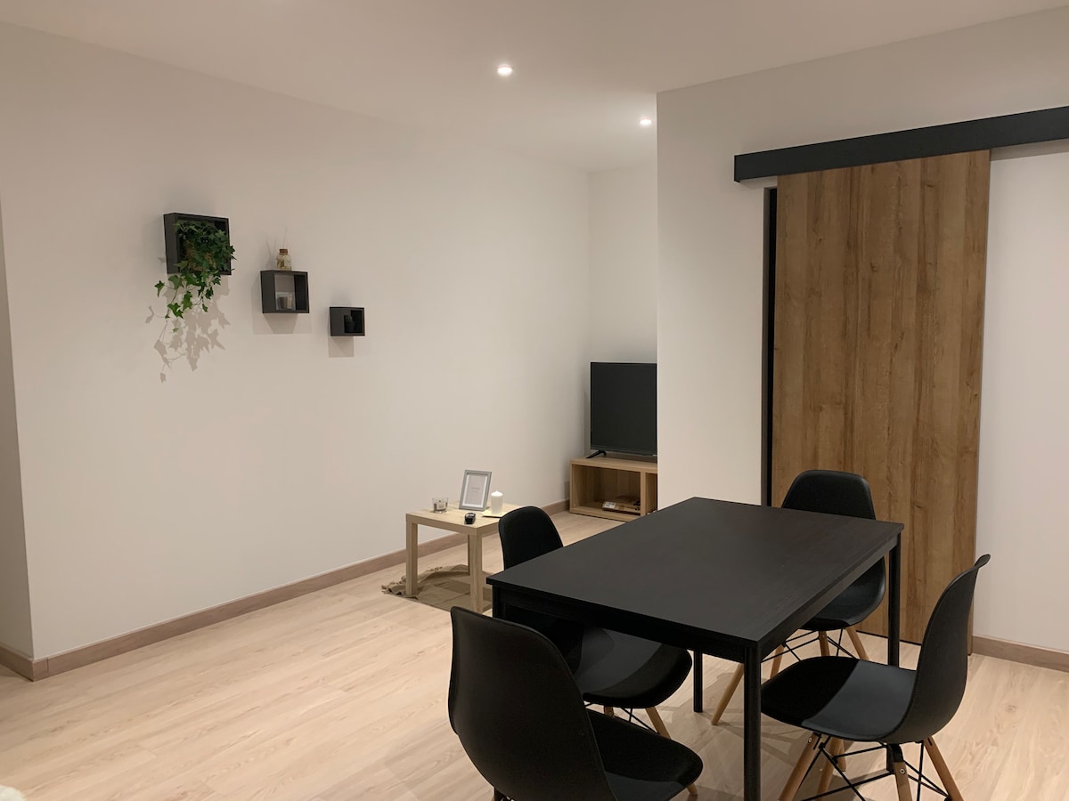 A spacious living area is shown, featuring a simple wooden dining table surrounded by four black chairs. A contemporary wall unit with decorative plants and framed pieces is visible, along with a flat-screen television on a low wooden stand. A sliding door opens to another space.