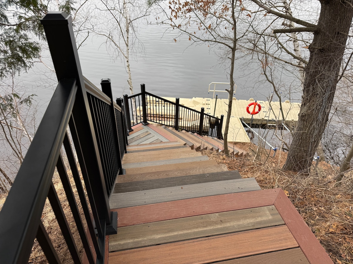 A winding staircase is shown, leading down to the water's edge. Each step is crafted from colorful, slip-resistant decking materials. On the left, trees frame the view, and the calm water reflects the surrounding nature, with a dock visible at the bottom of the stairs.