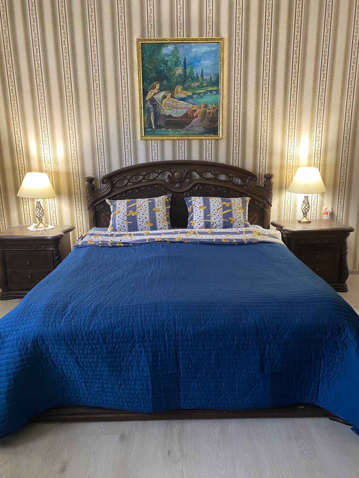 A cozy bedroom is furnished with a wooden bed featuring intricate headboard details. The bed is dressed in a blue quilt and accented by patterned pillows. Artwork hangs on the wall, complemented by soft bedside lamps that provide gentle lighting.