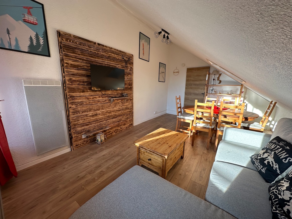 The spacious living area features a large wooden television unit against a sloped wall. A light-colored sofa is positioned nearby, with a wooden coffee table in front. A dining table surrounded by wooden chairs is visible in the background, illuminated by natural light from a window.