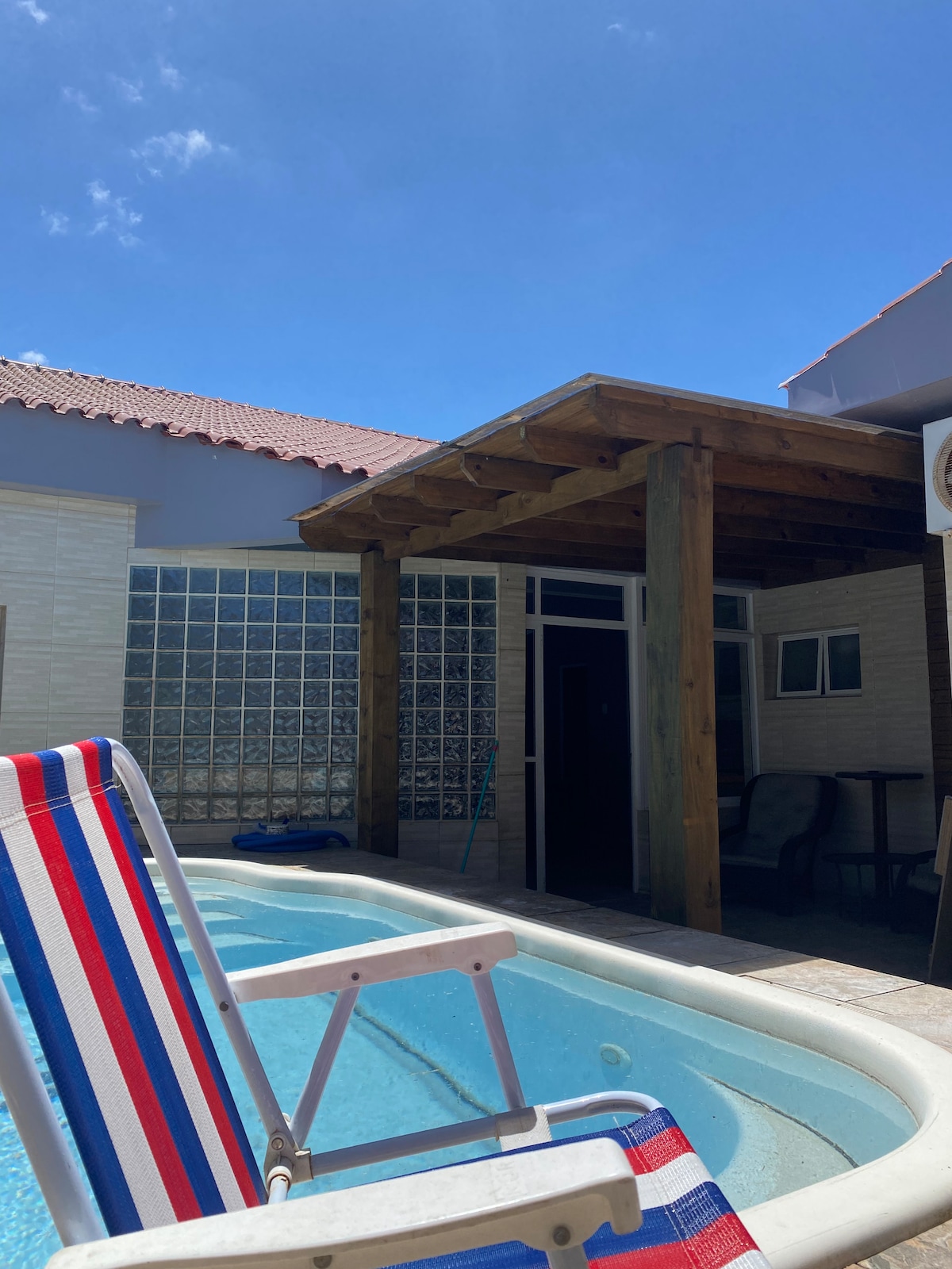 A sunny outdoor space is showcased, featuring a private pool with a wooden pergola nearby. A striped lounge chair is positioned beside the pool, offering a relaxing place to enjoy the surroundings. The backdrop consists of a glass block wall and a partially shaded area.