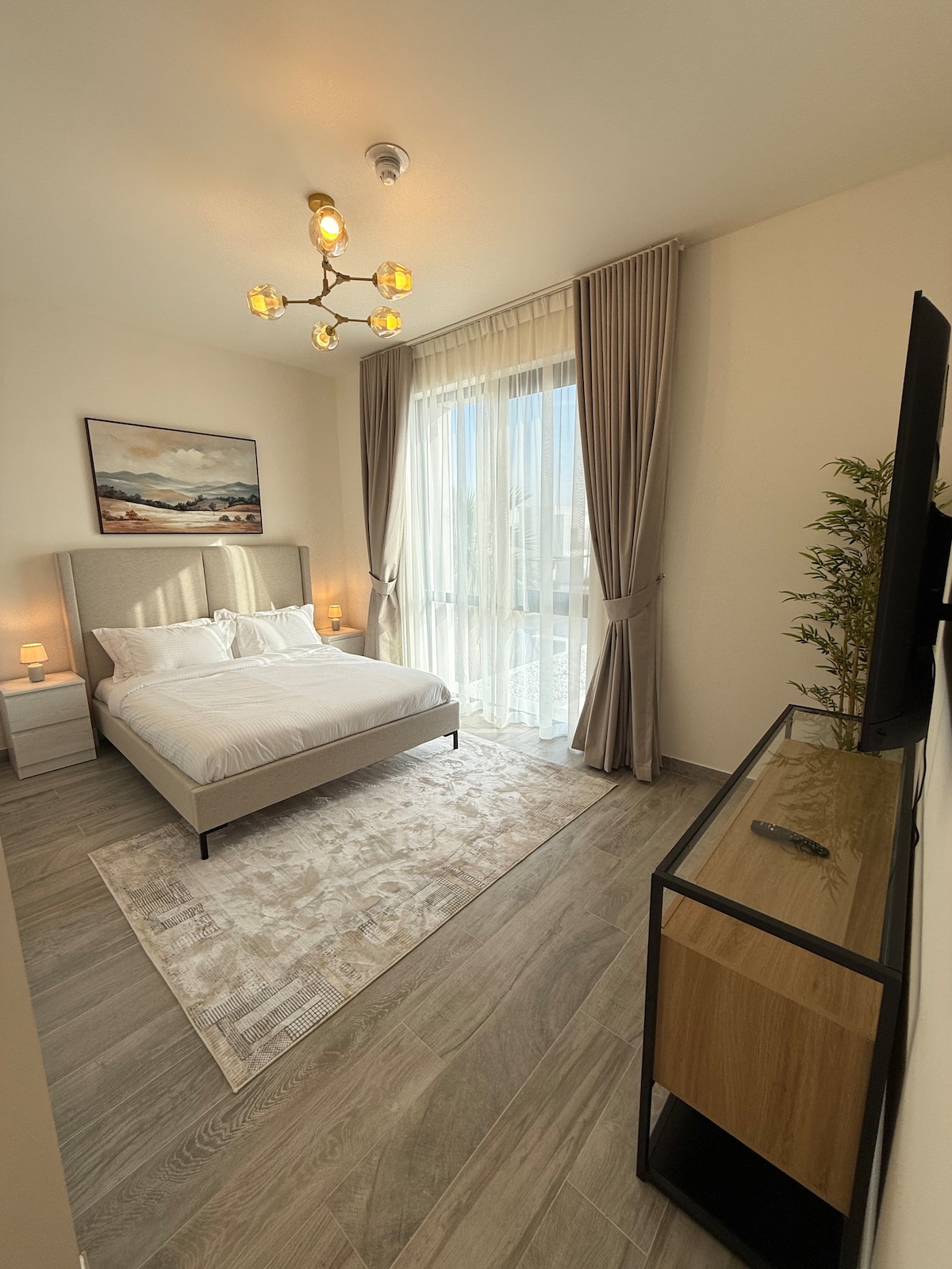 A light-filled bedroom features a plush bed with crisp linens and decorative pillows. Large windows draped with soft curtains allow natural light to fill the space, while a stylish area rug adds warmth. A modern console with a television is positioned near the entrance.