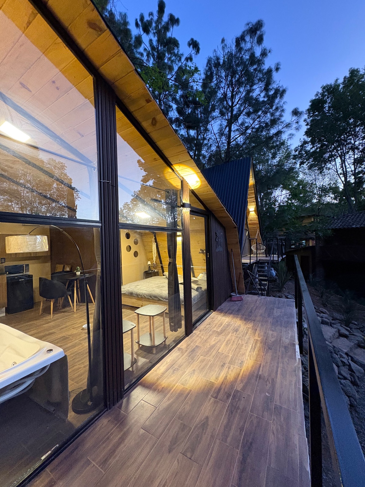 A wooden cabin is depicted at dusk, showcasing large glass windows that offer a view of the interior. A spacious deck extends along the side, while the environment is surrounded by trees, creating a serene atmosphere. Outdoor lighting softly illuminates the facade.
