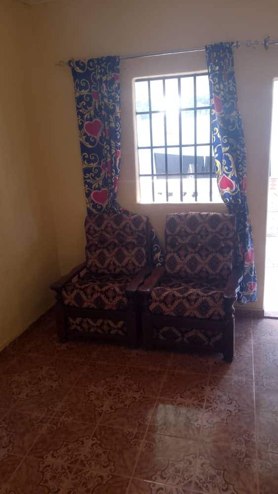 Two patterned wooden chairs are arranged near a window framed with colorful curtains. Natural light filters through the window, highlighting the textured floor tiles and the warm, yellow walls of the room.