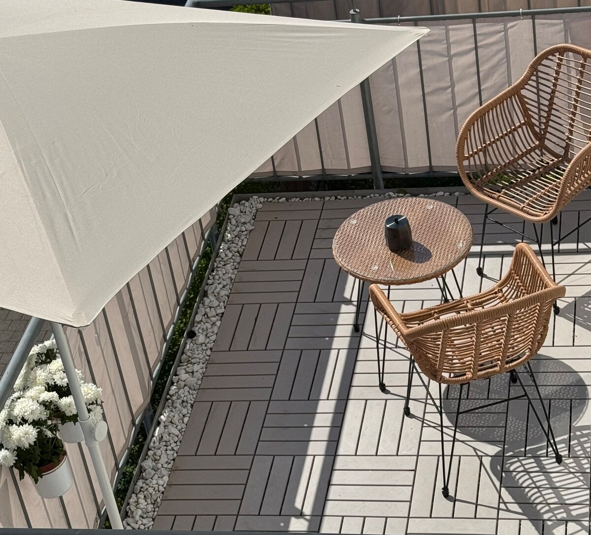 A small balcony is furnished with two rattan chairs and a round table, providing a cozy space for relaxation. A neutral-colored sunshade offers protection from sunlight, while a planter with white flowers adds a touch of nature. The curved lines of the furniture create a welcoming atmosphere.
