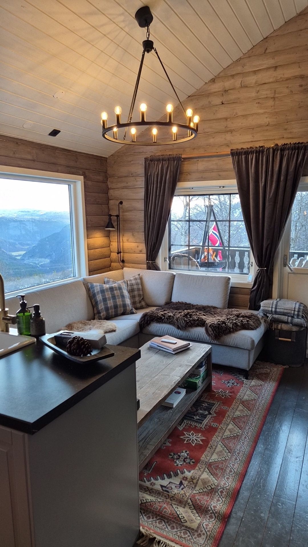 A cozy seating area features a light-colored sectional couch adorned with checkered pillows and a soft blanket. A wooden coffee table sits in front, while large windows frame views of the surrounding landscape, enhanced by natural light. Elegant curtains add a touch of warmth.