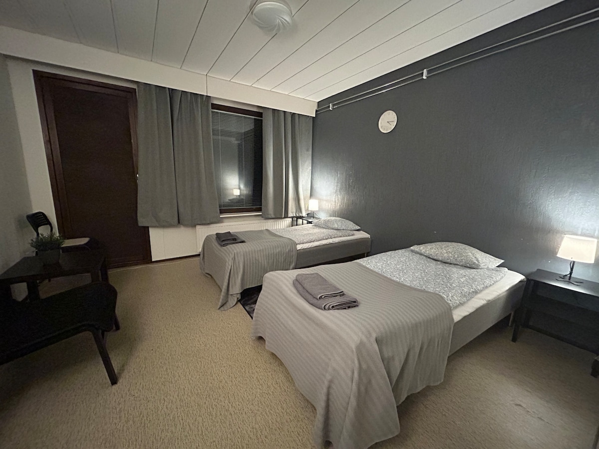 A comfortably arranged sleeping area features two spacious beds with light bedding. A side table with a lamp is positioned next to each bed. Soft curtains frame a large window, while a small desk and chair are located in the corner.