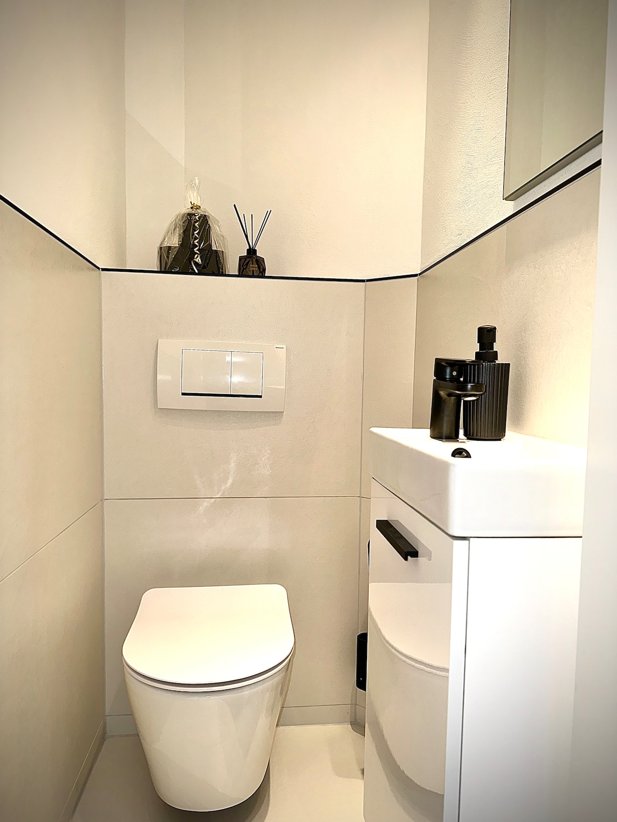 A modern bathroom features a sleek, white toilet and a contemporary sink with a minimalist black faucet. Decorative items are positioned on a ledge above the toilet, and a mirror reflects the clean lines of the space. Neutral tones create a calming atmosphere.