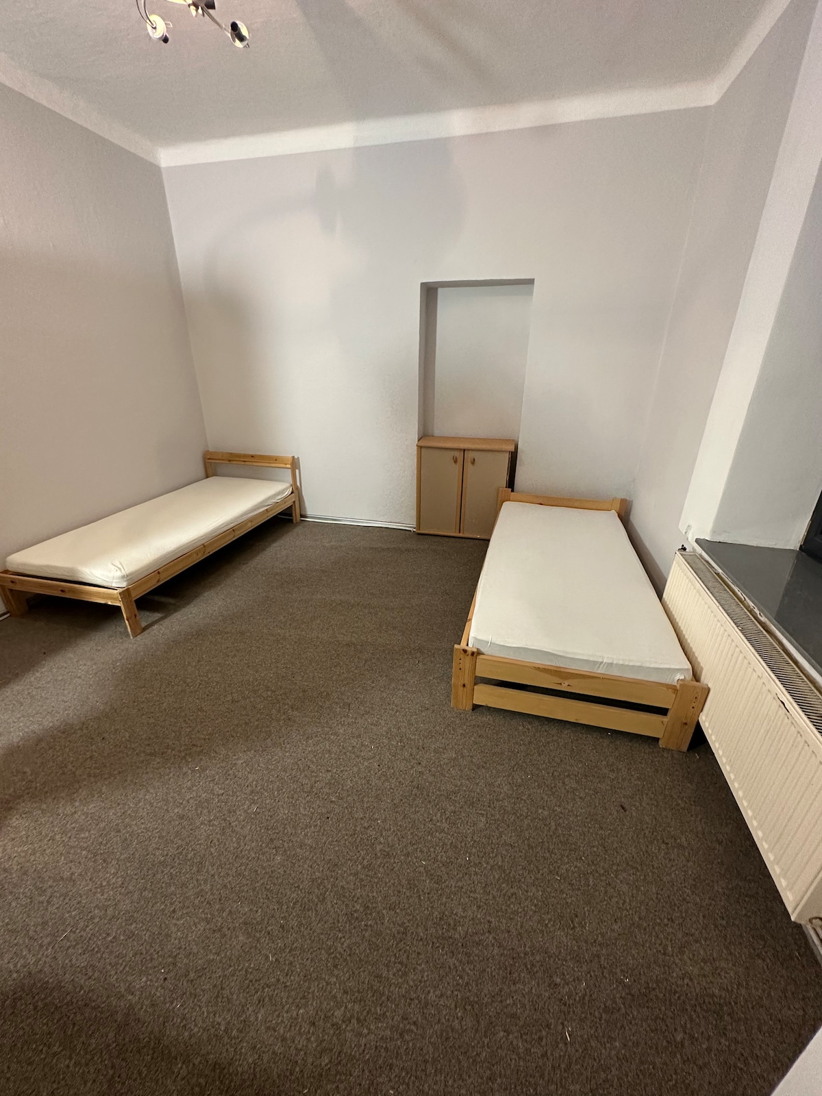 A spacious room features two wooden single beds positioned opposite each other. A small cabinet is located against the wall, with a nook in between the beds. The floor is covered with neutral-toned carpet, and soft lighting brightens the simple, clean space.