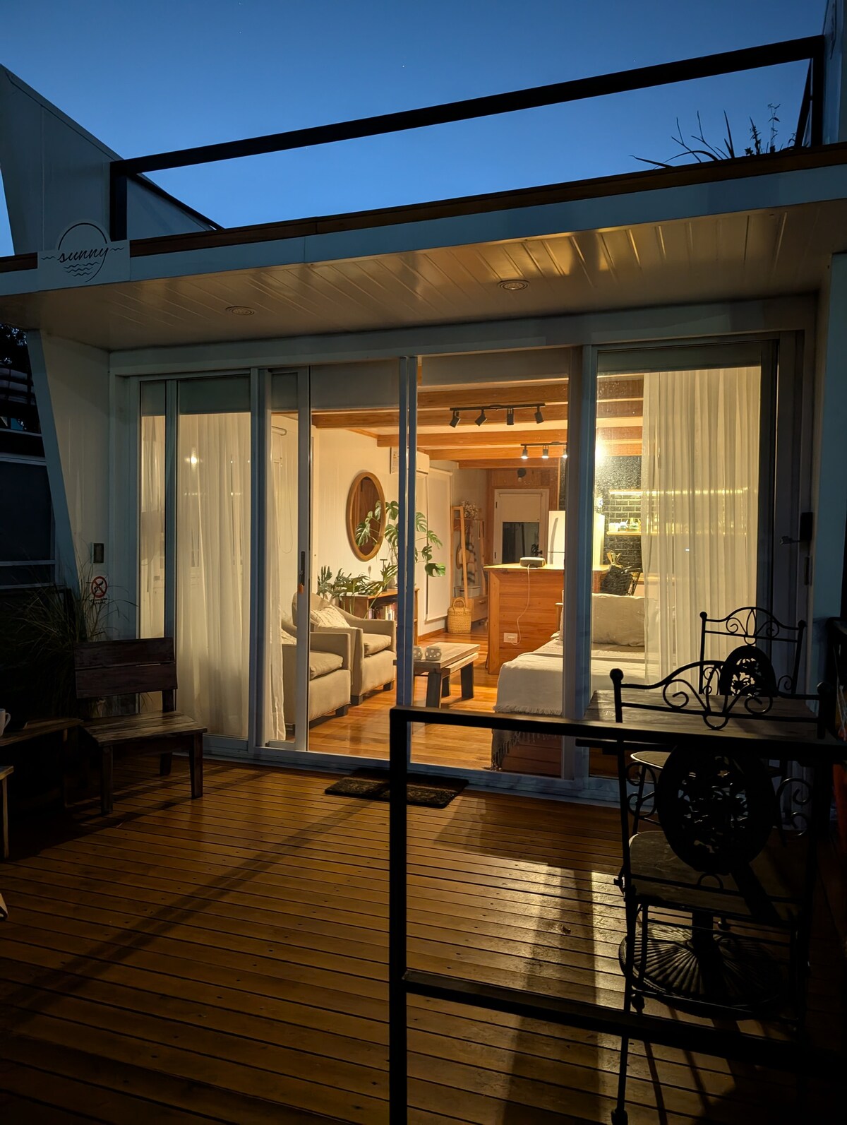 The exterior of the houseboat is captured at dusk, showcasing large glass sliding doors that provide a view into the warmly lit interior. A wooden deck leads to the entrance, surrounded by nearby seating arrangements, emphasizing a cozy atmosphere.