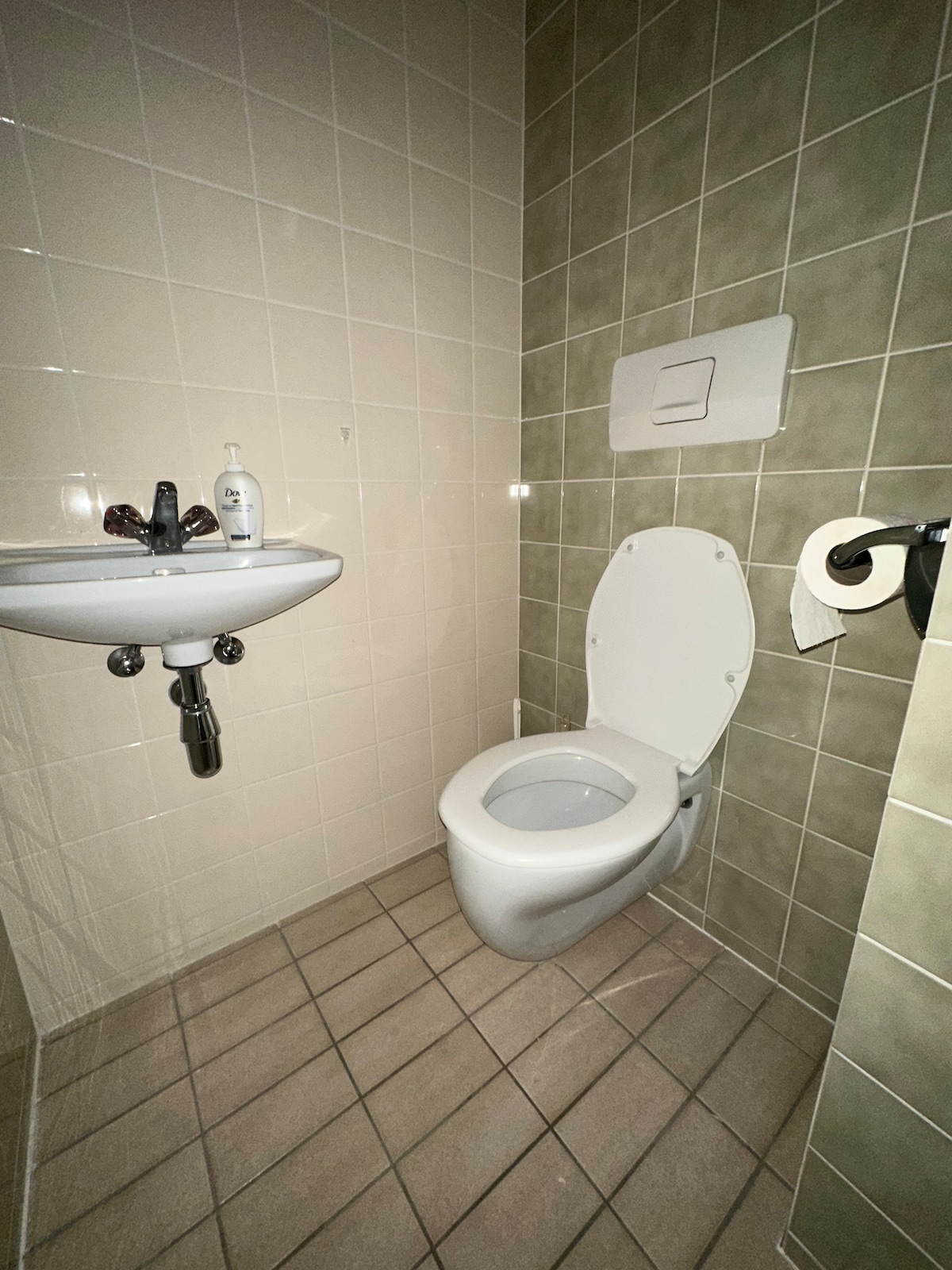 A modern bathroom featuring a clean and minimalistic design. A wall-mounted sink is positioned next to a toilet, both contributing to the functional layout. Neutral tiles cover the walls, while the floor is adorned with rectangular tiles in a muted tone.