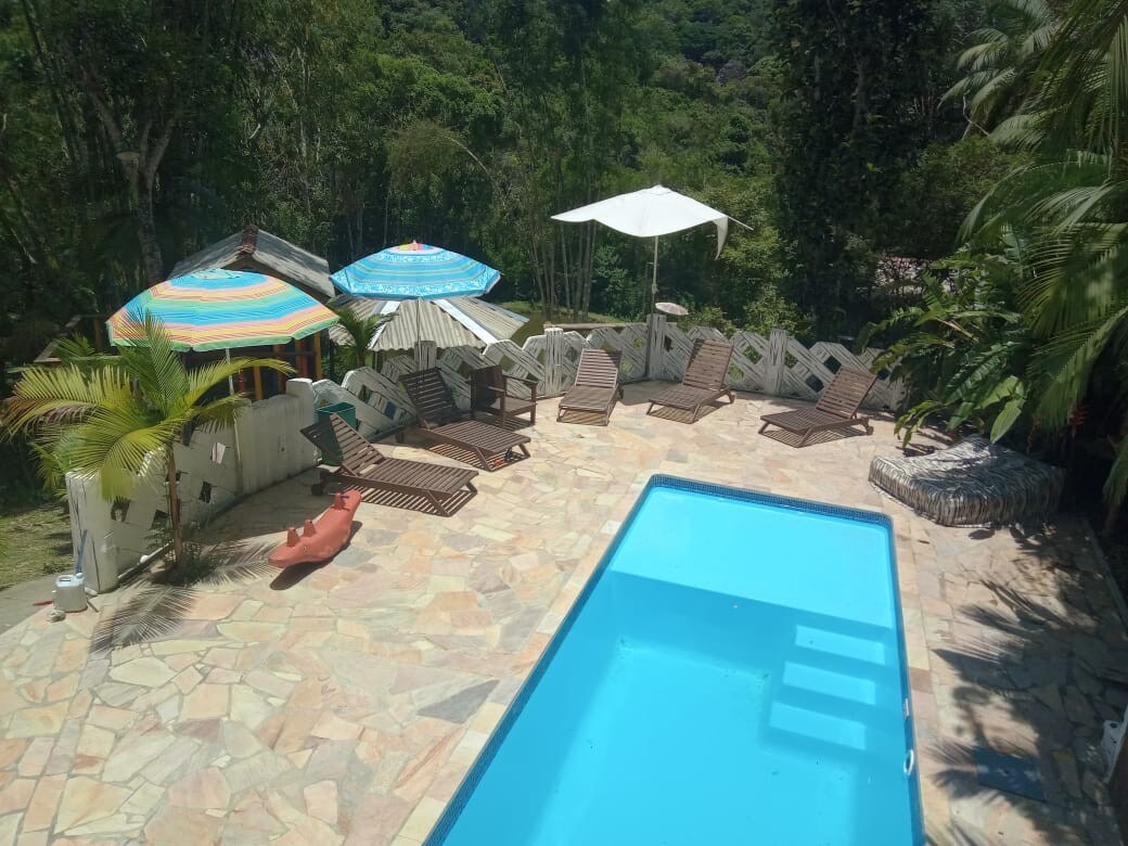 A refreshing swimming pool is surrounded by sun loungers and colorful umbrellas, providing an inviting space for relaxation. Lush greenery is visible in the background, enhancing the serene atmosphere of the outdoor area.