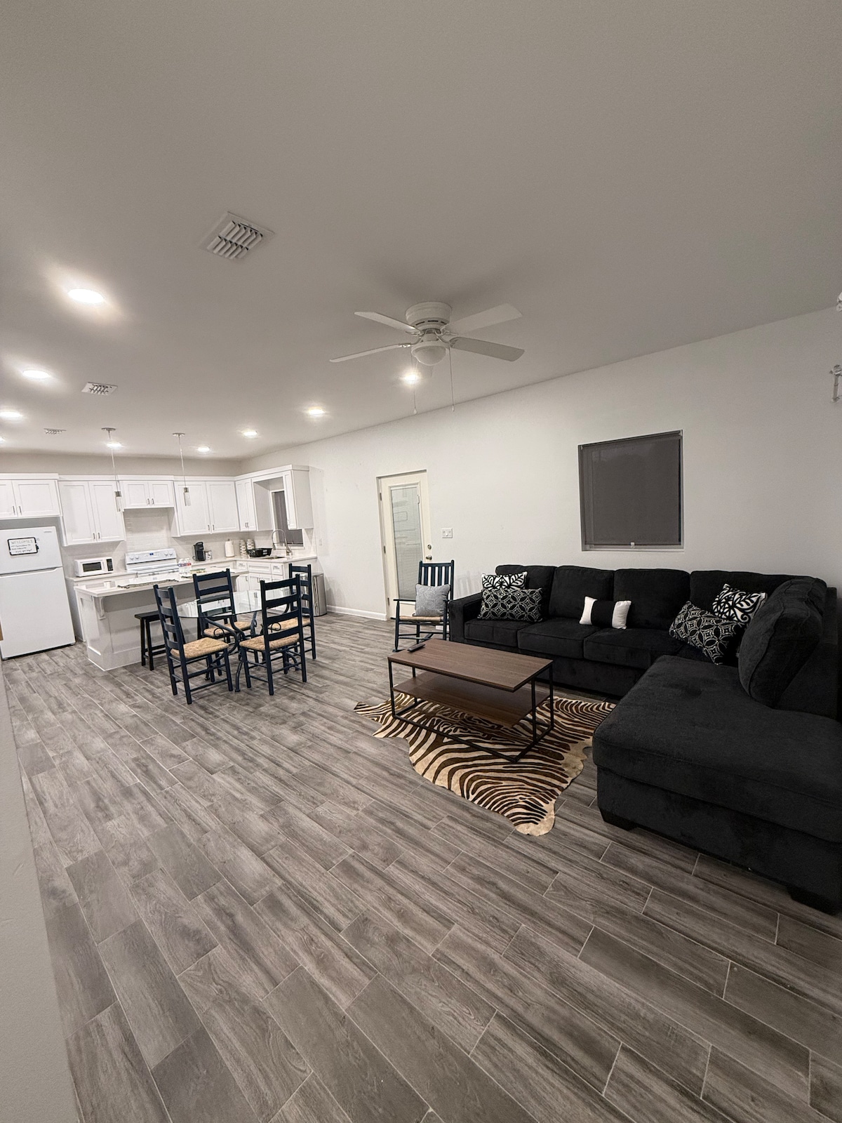 A living area is furnished with a sectional sofa and a coffee table atop a patterned rug. A dining area with a set of dark chairs and a table is visible. The kitchen is open and equipped with white cabinetry and appliances, with light coming from overhead fixtures.