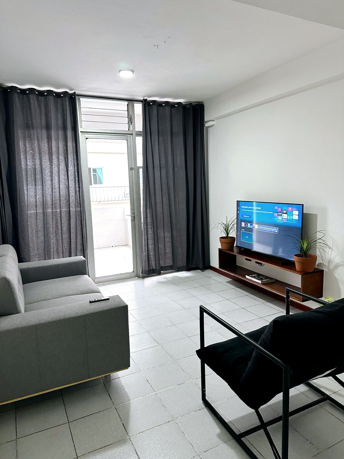 A living area is presented with a comfortable gray sofa and a black chair facing a wall-mounted 50-inch TV. Natural light enters through a balcony door, partially draped with dark curtains. Small potted plants add a touch of greenery.