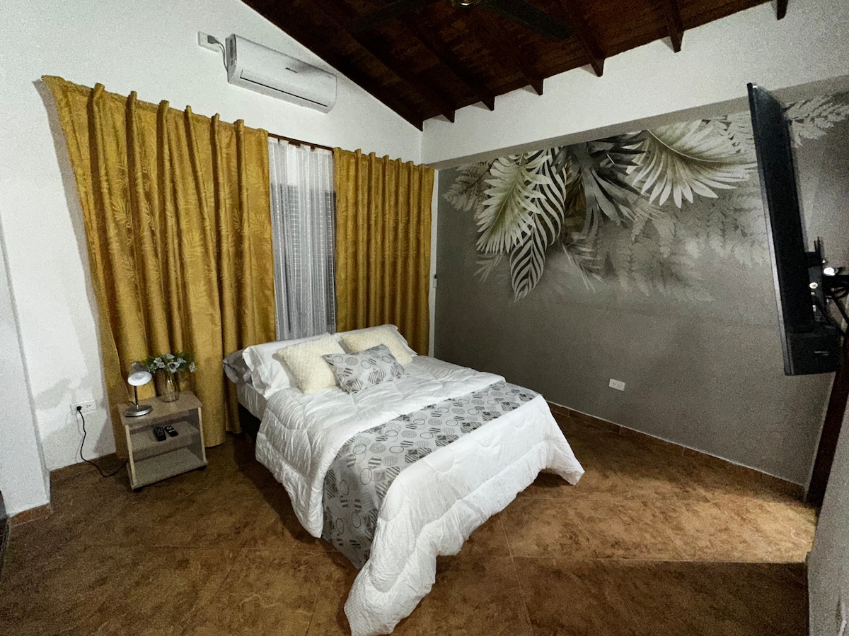 The sleeping area features a spacious bed with a light-colored comforter and decorative pillows. Golden curtains frame the window, while a mural of leaves adorns the adjacent wall. A bedside table holds small decorative items, complemented by warm, natural tones throughout the room.