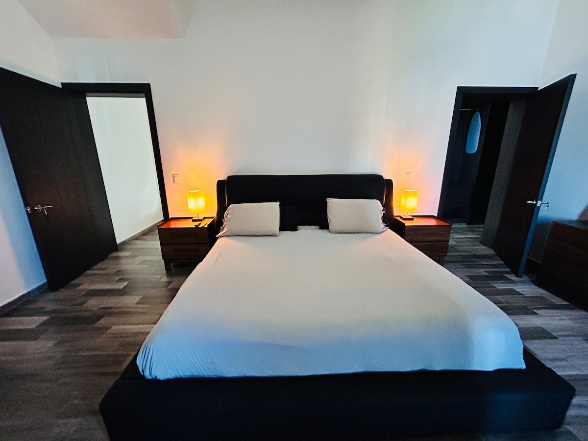 A spacious bedroom is observed, featuring a large bed with a neutral-colored bedding. Nightstands with lamps are positioned on either side of the bed, casting a soft glow. The room has dark wooden doors and contemporary flooring, contributing to its modern aesthetic.