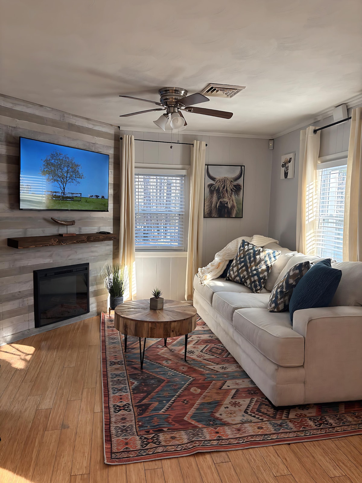 A bright living area features a light-colored sofa adorned with patterned cushions. A round wooden coffee table sits on a colorful area rug, and a wall-mounted television displays a scenic landscape. Sunlight enters through partially open window treatments, complementing the room's neutral tones.