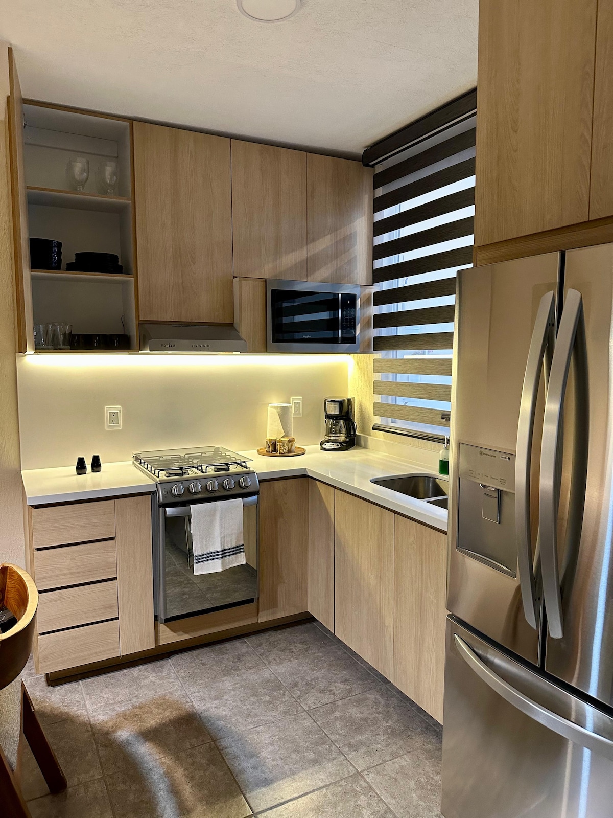 A modern kitchen is displayed with wood cabinetry and white countertops. Stainless steel appliances, including a refrigerator and stove, are visible. Soft, ambient lighting highlights an organized countertop with a coffee maker. Horizontal blinds allow filtered light to enter the space, creating a bright atmosphere.