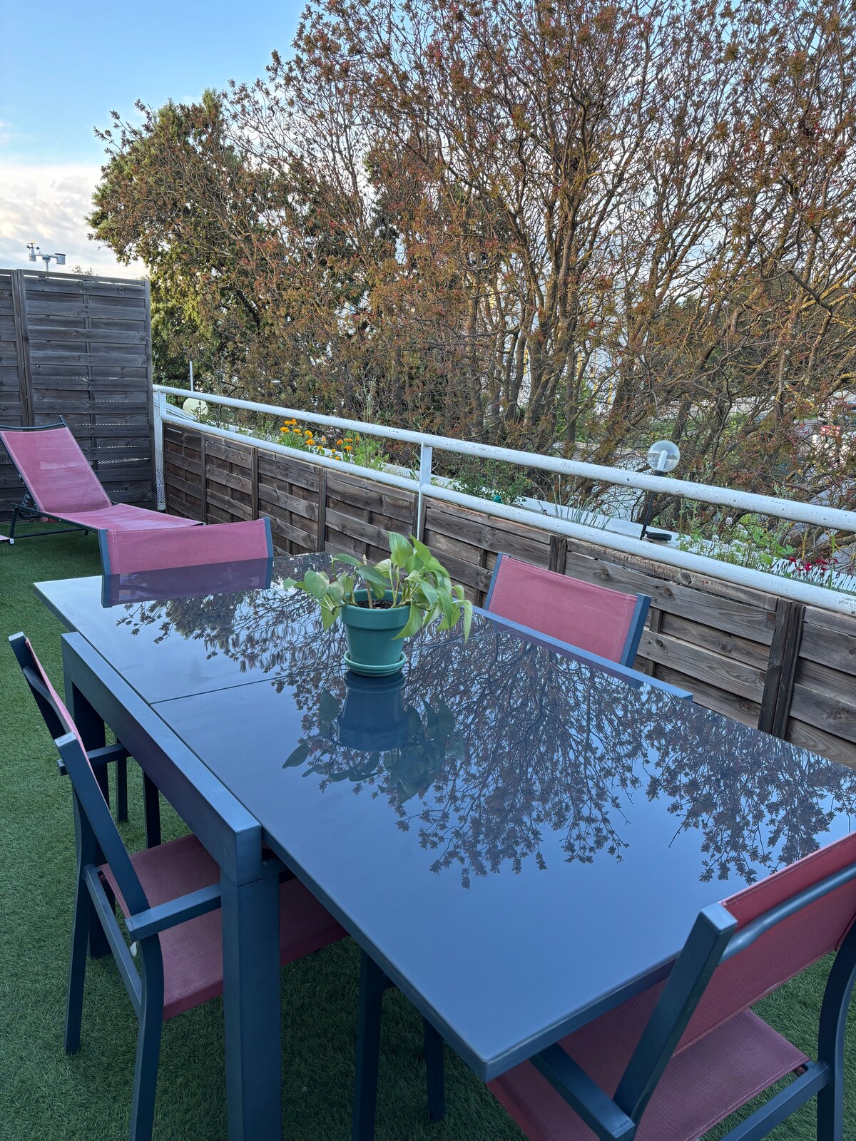 A spacious terrace features a dining table with a glossy surface, surrounded by six red chairs. Potted plants are visible on the table, with sunlight reflecting off the furniture. Lush greenery lines the perimeter, offering a serene outdoor setting.