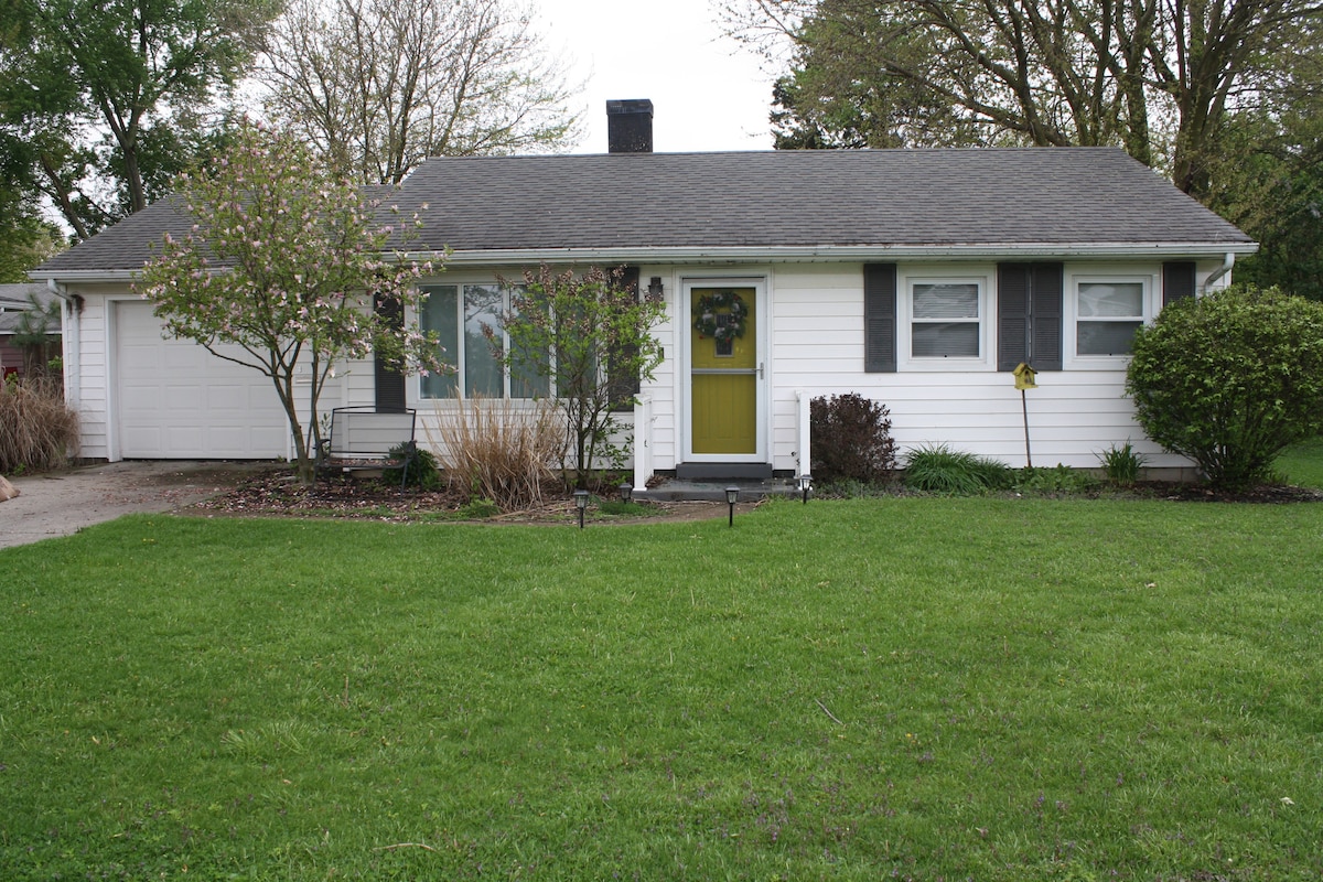 A single-story home is presented, featuring a light exterior with gray shutters and a welcoming yellow front door. A manicured lawn surrounds the house, complemented by small shrubs and blossoming trees on either side. A garage is visible on the left side of the property.