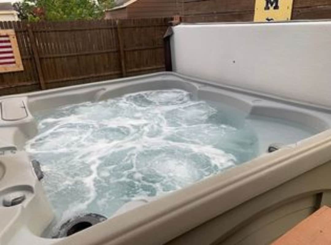 A hot tub is seen with bubbling water, situated within a fenced outdoor area. The edges are framed by light-colored siding and wooden decking, creating a serene space for relaxation. The background features a wooden fence adding to the privacy of the area.