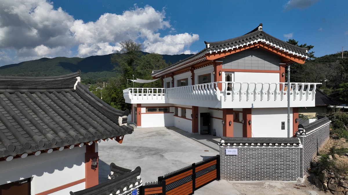 A traditional-style building is shown with a distinctive tiled roof and ornamental eaves, set against a backdrop of rolling mountains. The spacious exterior includes a large balcony, and a well-defined courtyard area can be seen, with ample parking space available.